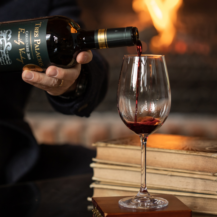 Pouring Tres Palacios Carmenere 2019 into a glass in a cozy setting with a warm fireplace and vintage books, highlighting a relaxed wine experience.