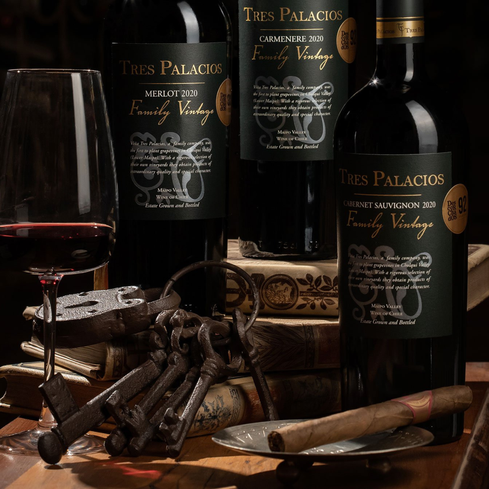 Three bottles of Tres Palacios wine (Merlot, Carmenere, and Cabernet Sauvignon 2020), paired with a glass of red wine, antique keys, and books, creating a luxurious, refined atmosphere.