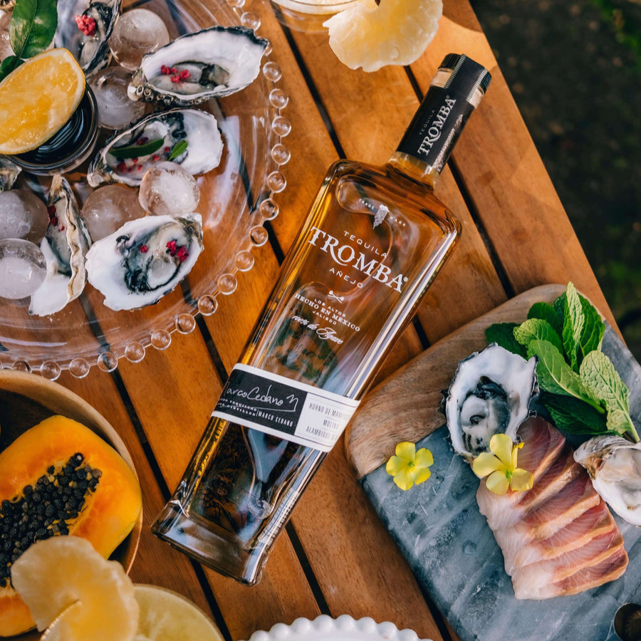 Tromba Añejo Tequila bottle beautifully styled with fresh oysters and tropical fruits on a wooden table.