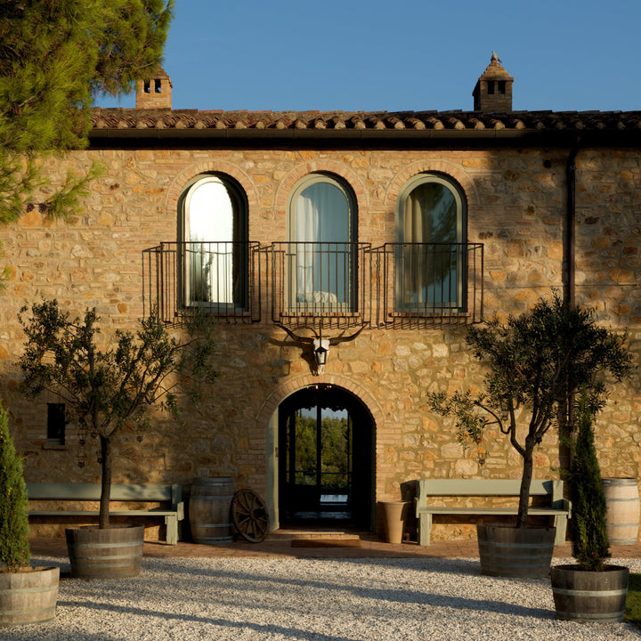 A Tuscan villa with a stone facade, arched windows, a balcony, and a decorative bull skull above the entrance. Olive trees and benches flank the entrance.