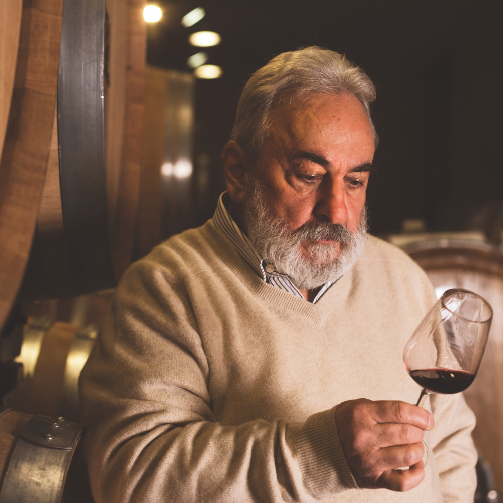 A Vinchio Vaglio winemaker carefully evaluating red wine in a glass surrounded by barrels in a cellar.