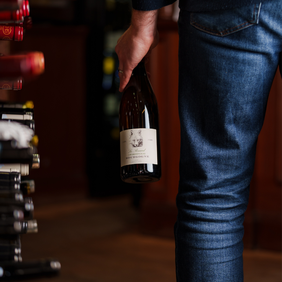 A person holding a bottle of Bourgogne Chardonnay in a wine shop, standing near a shelf of various wine bottles, emphasizing a cozy, upscale ambiance.