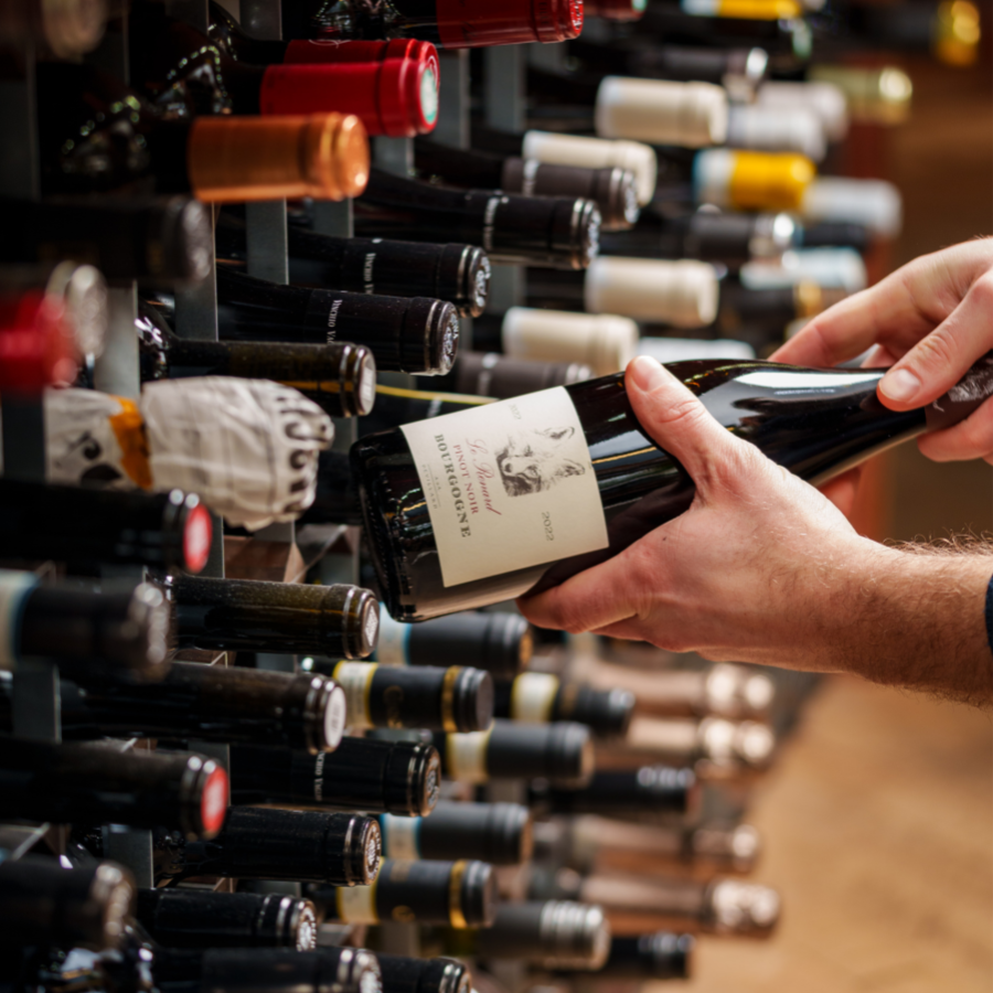 Hands picking a bottle of Bourgogne Pinot Noir 2020 from a wine rack in a store, surrounded by rows of various wine bottles, highlighting an elegant selection atmosphere.