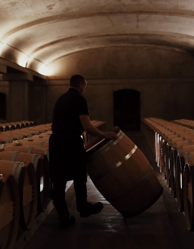 Winemaker rolling a wooden wine barrel in a dimly lit wine cellar.