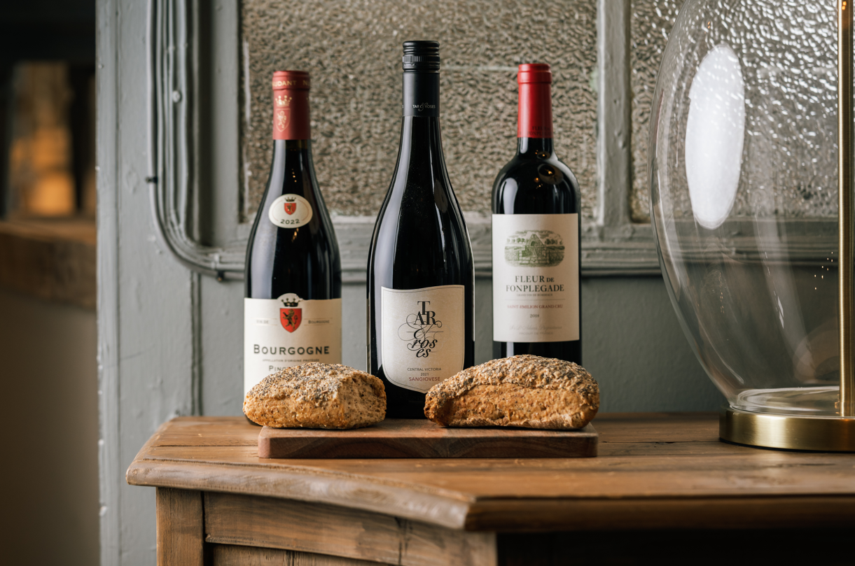 Three bottles of wine – a Bourgogne Pinot Noir 2022, a Central Victoria Sangiovese, and a Fleur de Fonplegade Saint-Émilion Grand Cru 2016 – displayed on a rustic wooden table with two artisanal bread loaves. A stylish setting with textured glass and elegant décor.